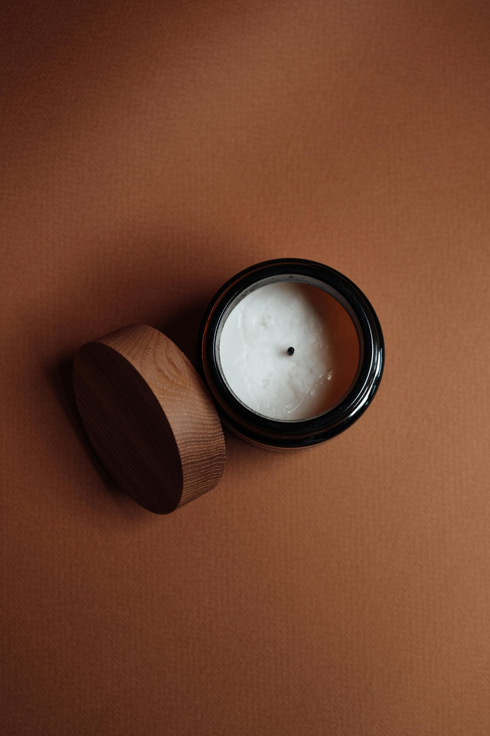 Top view of a candle in a jar with a wooden lid on a textured brown background.