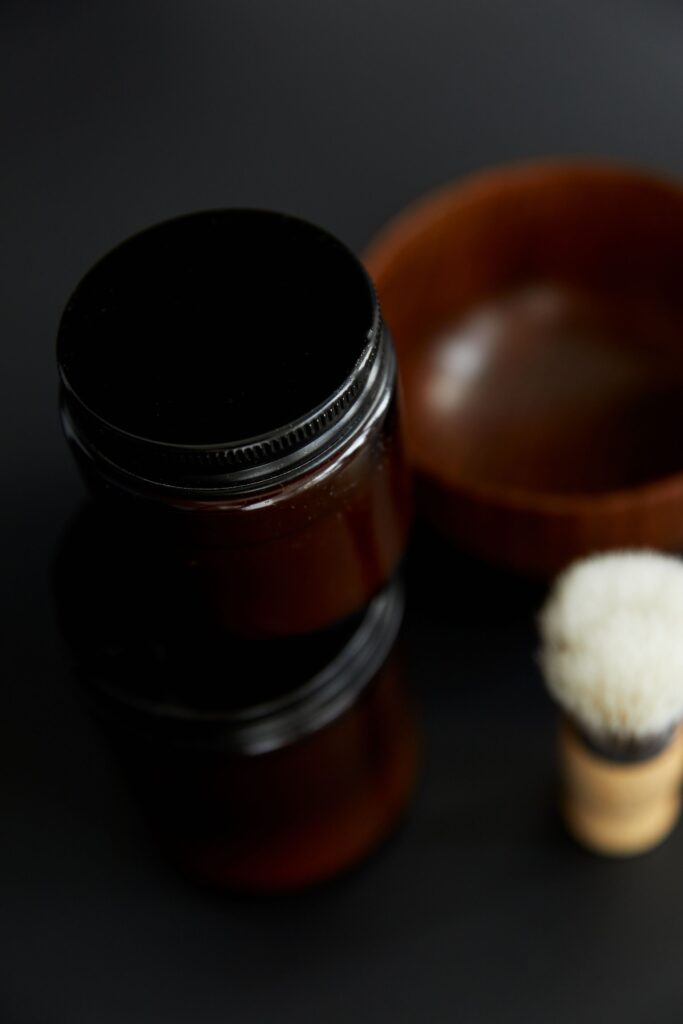 Close-up of a dark glass jar, wooden bowl, and shaving brush on a black surface, emphasizing grooming essentials.